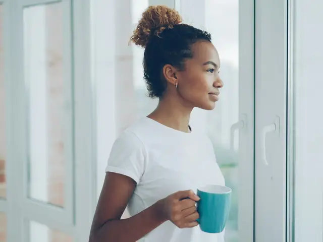 Eine junge Frau steht mit einer Tasse in der Hand am Fenster und schaut nachdenklich nach draußen.